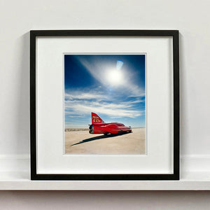 Black framed photograph by Richard Heeps. A red drag car with a 75 written on its fin sits on a salt plain the front facing away towards the right.  A blue cloudy sky is overhead.