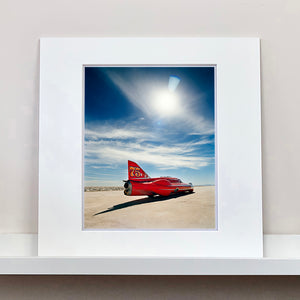 Mounted photograph by Richard Heeps. A red drag car with a 75 written on its fin sits on a salt plain the front facing away towards the right. A blue cloudy sky is overhead.