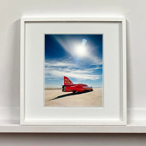 White framed photograph by Richard Heeps. A red drag car with a 75 written on its fin sits on a salt plain the front facing away towards the right. A blue cloudy sky is overhead.