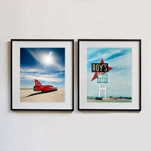 Two black framed photographs by Richard Heeps. The left hand side photograph is of a red drag car with a 75 written on its fin sits on a salt plain the front facing away towards the right. A blue cloudy sky is overhead. The photograph on the right hand side is a large sign for Roy's Motel and Cafe, on flat land with mountains in the background and a vast sky behind.. 