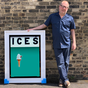 Photograph by Richard Heeps.  Richard Heeps holds a white framed print. At the top of the print, black letters spell out ICES and below is depicted a 99 icecream cone sitting left of centre against a Turquoise Teal coloured background.  