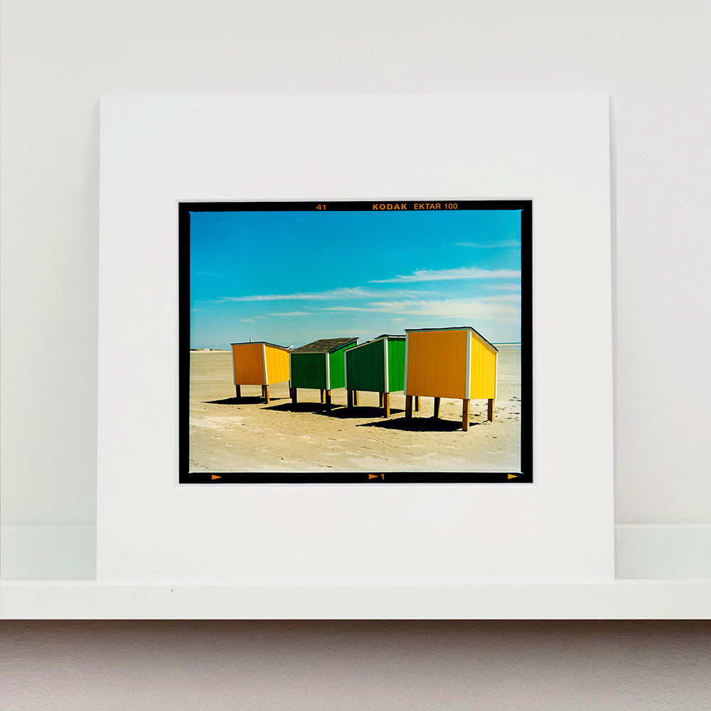 Mounted photograph by Richard Heeps. Yellow and green beach lockers stand on their wooden legs on the clear sandy beach.