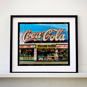 Black framed photograph by Richard Heeps. Harry's Corner was taken on the wildwood boardwalk, featuring neon typography and the iconic Coca-Cola sign against a bright blue sky. 