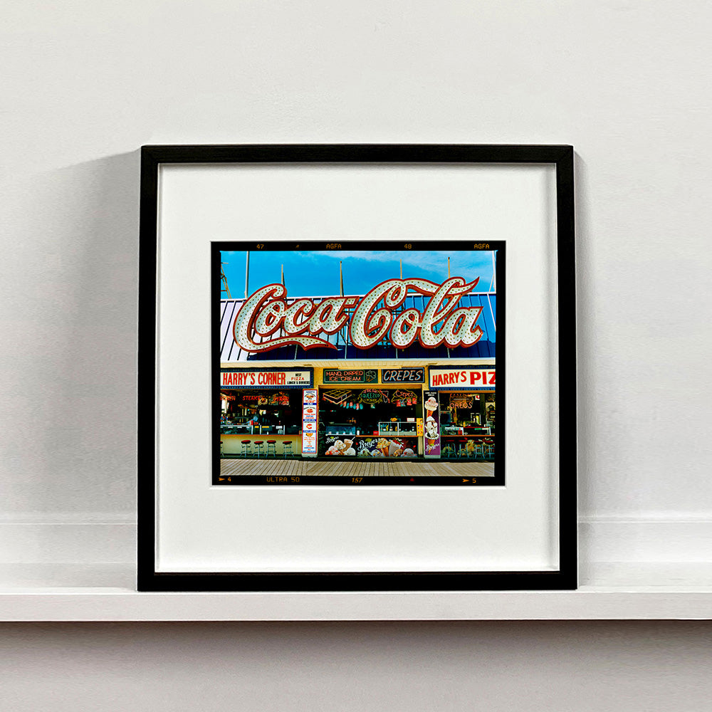 Black framed photograph by Richard Heeps. Harry's Corner was taken on the wildwood boardwalk, featuring neon typography and the iconic Coca-Cola sign against a bright blue sky. 