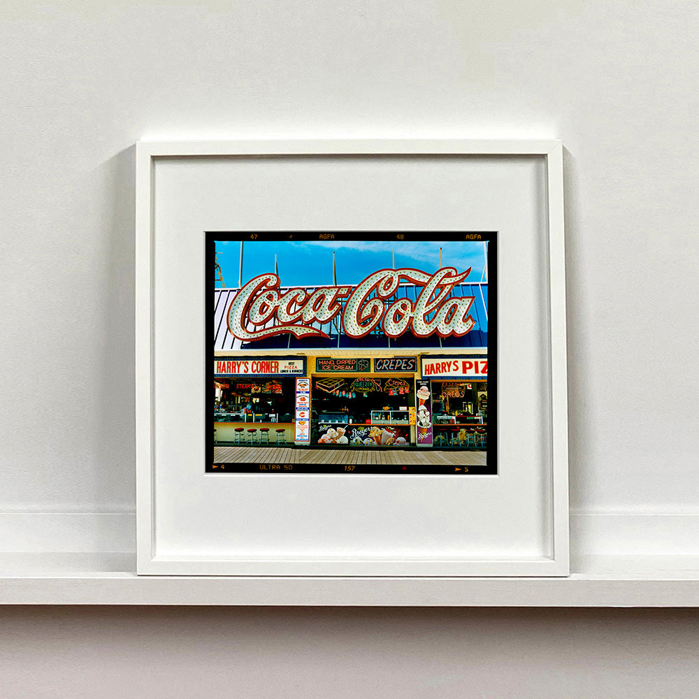 White framed photograph by Richard Heeps. Harry's Corner was taken on the wildwood boardwalk, featuring neon typography and the iconic Coca-Cola sign against a bright blue sky. 