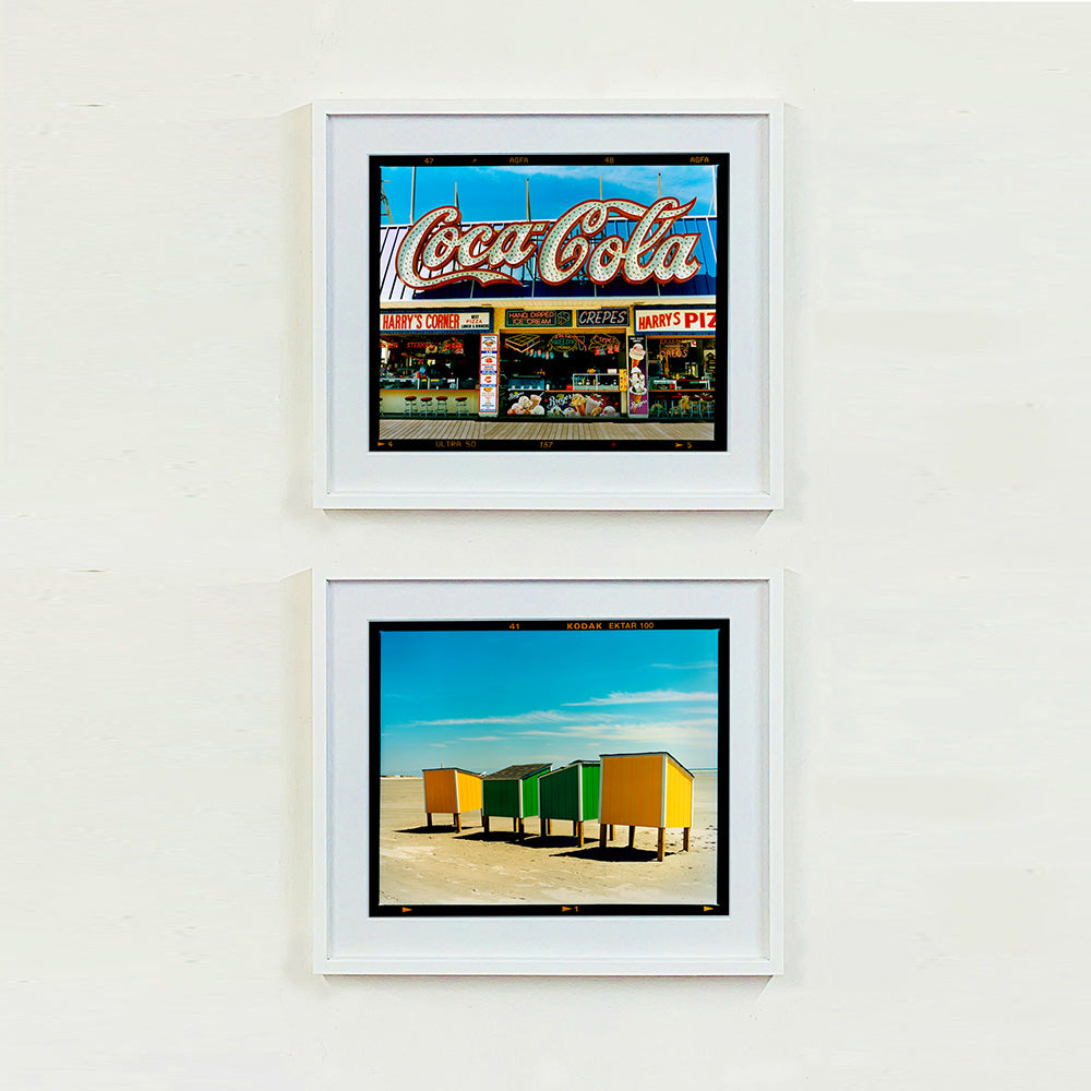 Two white framed photographs by Richard Heeps. The top photograph is of Harry's Corner and was taken on the wildwood boardwalk, featuring neon typography and the iconic Coca-Cola sign against a bright blue sky. The bottom photograph features four beach lockers, the two middle ones green and the two outer ones yellow, sitting on an empty beach with a blue sky.