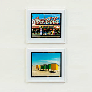 Two white framed photographs by Richard Heeps. The top photograph is of Harry's Corner and was taken on the wildwood boardwalk, featuring neon typography and the iconic Coca-Cola sign against a bright blue sky. The bottom photograph features four beach lockers, the two middle ones green and the two outer ones yellow, sitting on an empty beach with a blue sky.