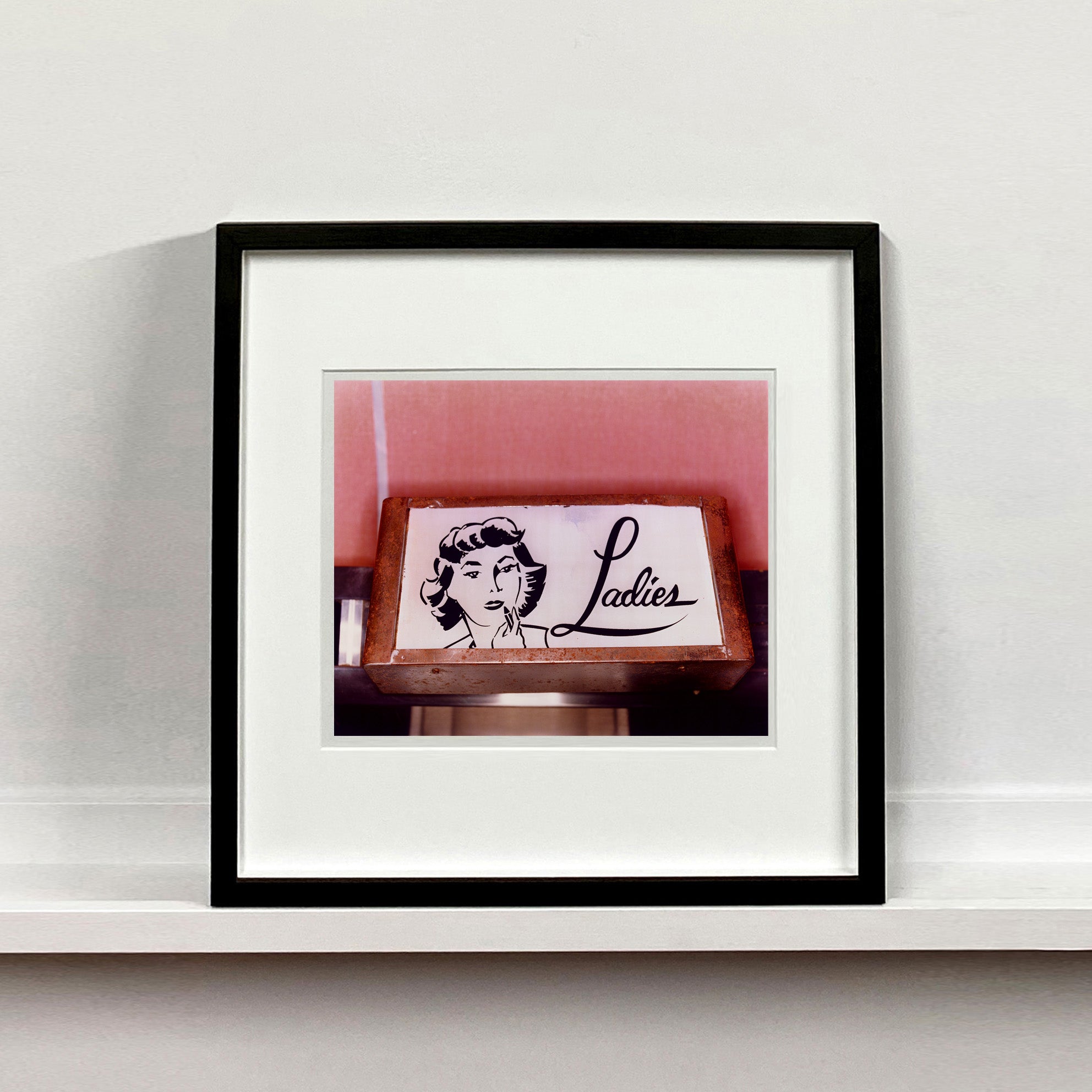 Black framed photograph by Richard Heeps. A kitsch Ladies' toilet sign. The sign has the word Ladies alongside an outline of 1950s woman. The sign sits in a wooden frame against a pink tiled wall.
