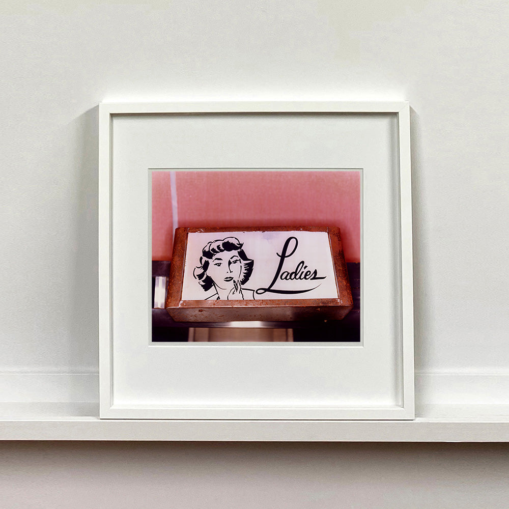White framed photograph by Richard Heeps. A kitsch Ladies' toilet sign. The sign has the word Ladies alongside an outline of 1950s woman. The sign sits in a wooden frame against a pink tiled wall.