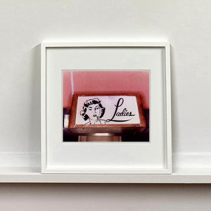 White framed photograph by Richard Heeps. A kitsch Ladies' toilet sign. The sign has the word Ladies alongside an outline of 1950s woman. The sign sits in a wooden frame against a pink tiled wall.