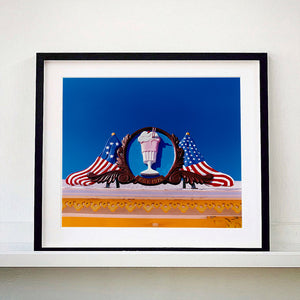 Black framed photograph by Richard Heeps. A 3D shape milkshake parlour sign which has a pink milkshake with a white top, cherry and straws, surrounded by a wooden type shield, and on either side the look of draped American flags. This is set against a blue sky.