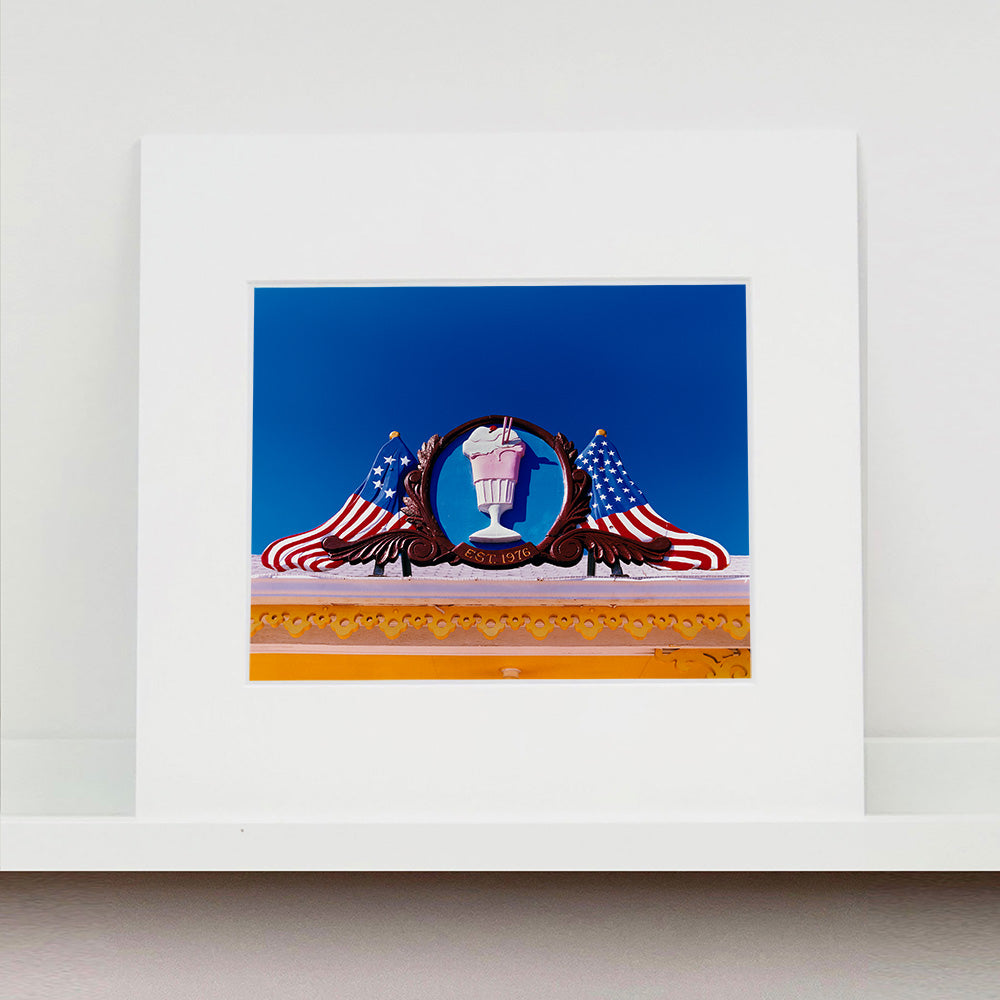 Mounted photograph by Richard Heeps. A 3D shape milkshake parlour sign which has a pink milkshake with a white top, cherry and straws, surrounded by a wooden type shield, and on either side the look of draped American flags. This is set against a blue sky.