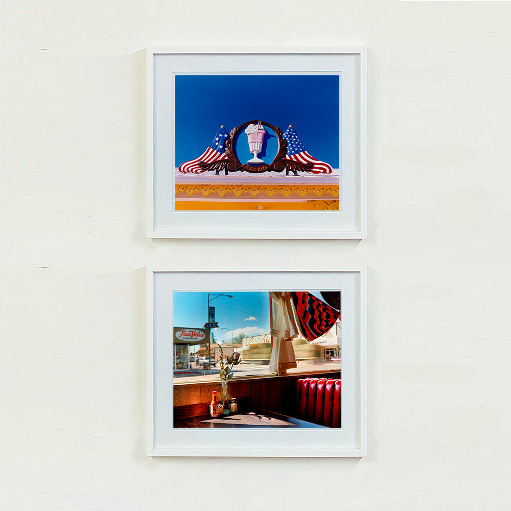 Two white framed photographs by Richard Heeps. The top photograph is of a 3D shape milkshake parlour sign which has a pink milkshake with a white top, cherry and straws, surrounded by a wooden type shield, and on either side the look of draped American flags. This is set against a blue sky. The bottom photograph is the inside of a retro diner with the classic red seating. The view is looking out of the window onto an American small town road.