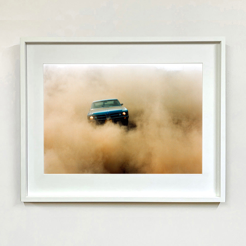 White framed photograph by Richard Heeps.  A light blue Buick car moving towards the camera and slightly obscured by the dust clouds which it has created.