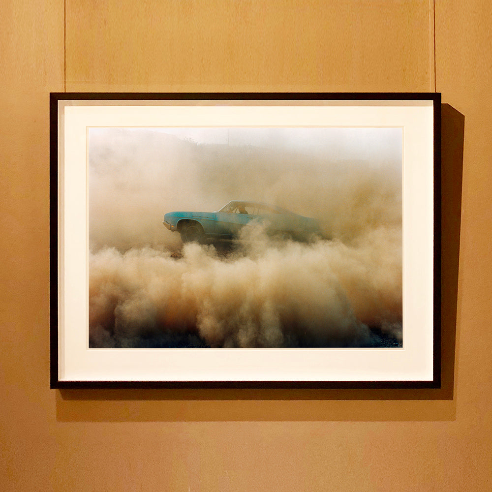 Black framed photograph by Richard Heeps. A side view of a light blue Buick car moving and slightly obscured by the dust clouds which it has created.