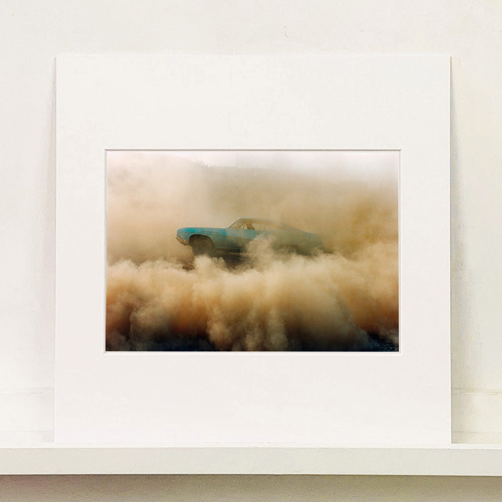 Mounted photograph by Richard Heeps. A side view of a light blue Buick car moving and slightly obscured by the dust clouds which it has created.