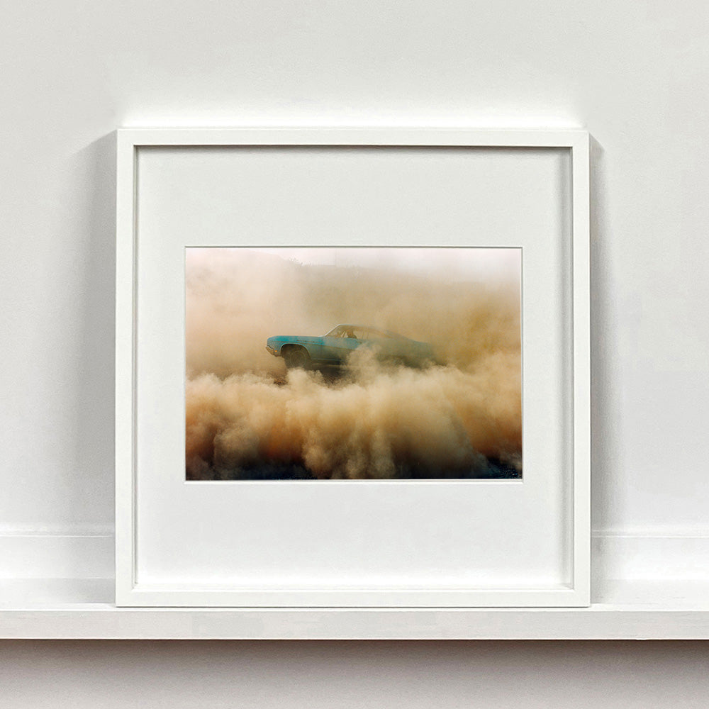 White framed photograph by Richard Heeps. A side view of a light blue Buick car moving and slightly obscured by the dust clouds which it has created.