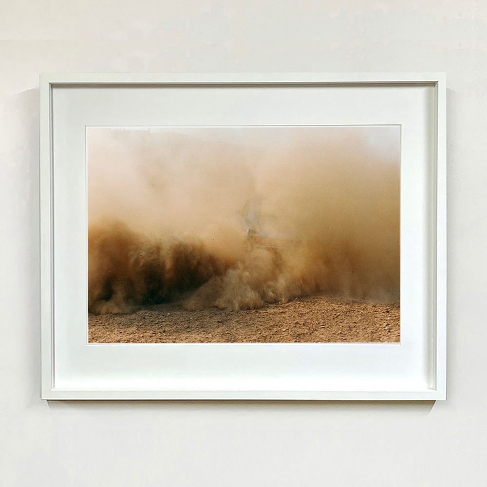 White framed mounted photograph by Richard Heeps. A vague outline of the back of a light blue Buick car moving and obscured by the dust clouds which it has created.