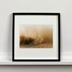Black framed mounted photograph by Richard Heeps. A vague outline of the back of a light blue Buick car moving and obscured by the dust clouds which it has created.