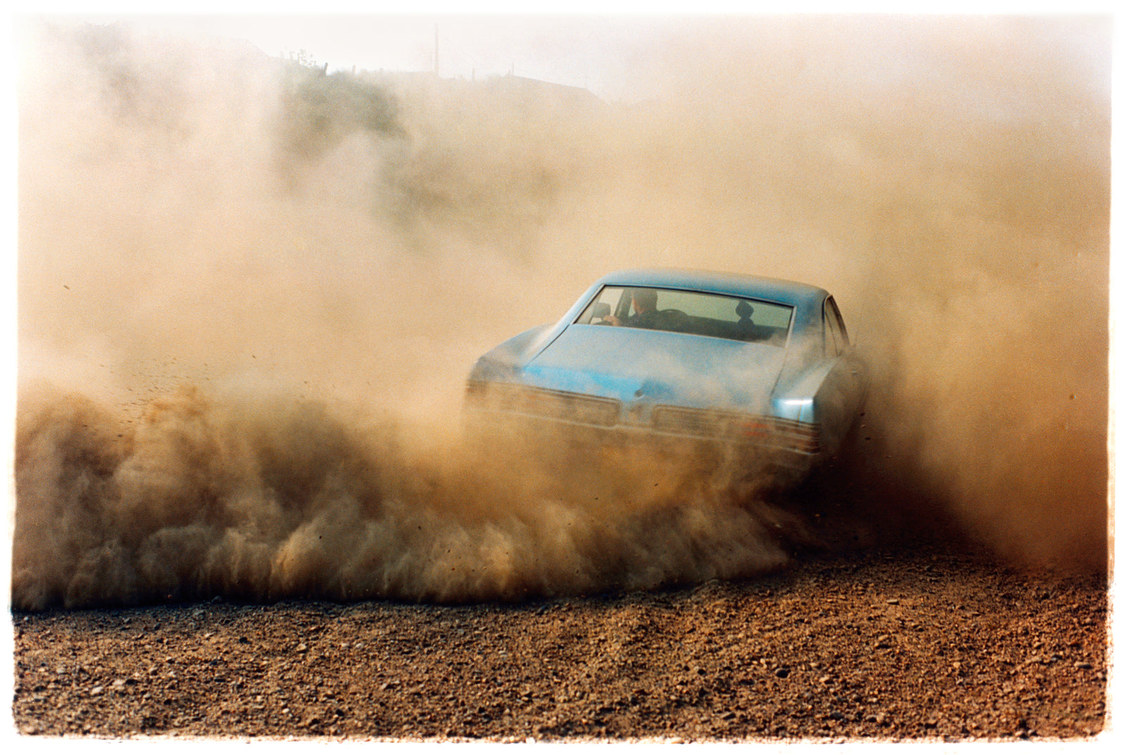 Photograph by Richard Heeps. A back view of a light blue Buick car moving and slightly obscured by the dust clouds which it has created.