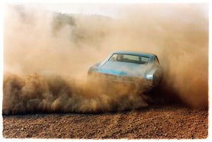 Photograph by Richard Heeps. A back view of a light blue Buick car moving and slightly obscured by the dust clouds which it has created.