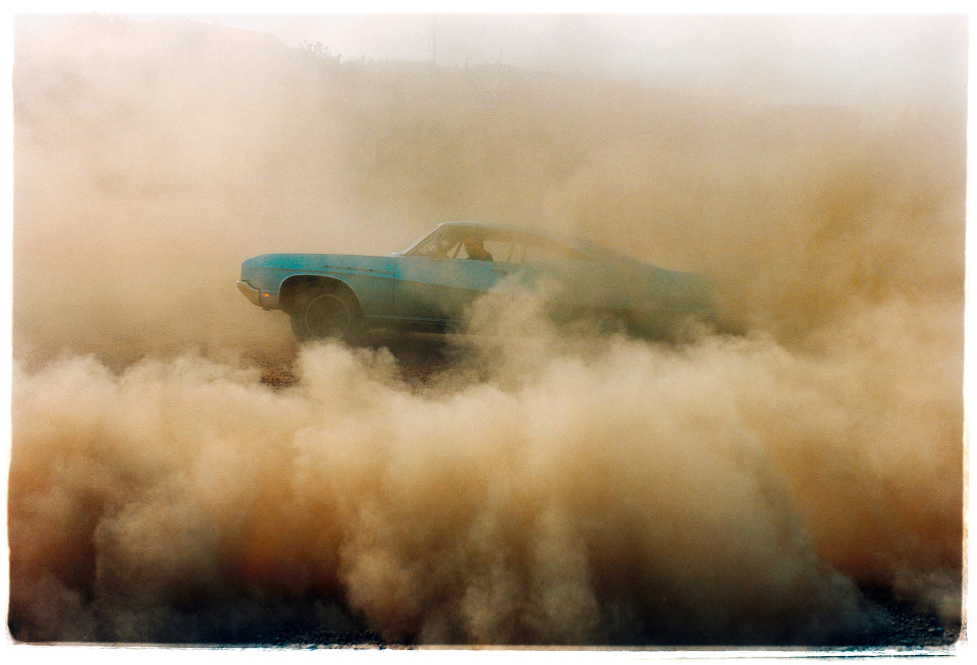 Photograph by Richard Heeps. A side view of a light blue Buick car moving and slightly obscured by the dust clouds which it has created.