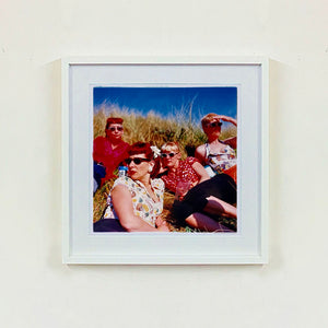 White framed photograph by Richard Heeps. A photo of four women dressed in a retro style all wearing sunglasses on a sunny day lounging on a hill with high grass.
