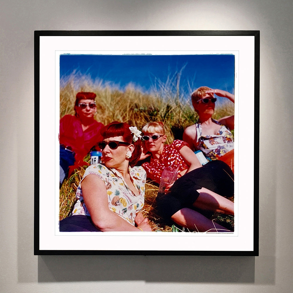 Black framed photograph by Richard Heeps. A photo of four women dressed in a retro style all wearing sunglasses on a sunny day lounging on a hill with high grass.