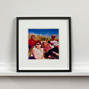 Black framed photograph by Richard Heeps. A photo of four women dressed in a retro style all wearing sunglasses on a sunny day lounging on a hill with high grass.
