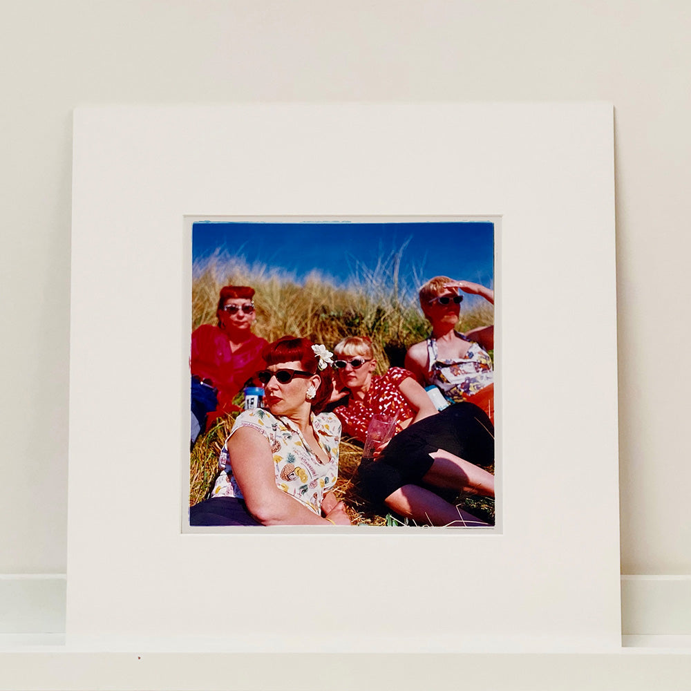 Mounted photograph by Richard Heeps. A photo of four women dressed in a retro style all wearing sunglasses on a sunny day lounging on a hill with high grass.