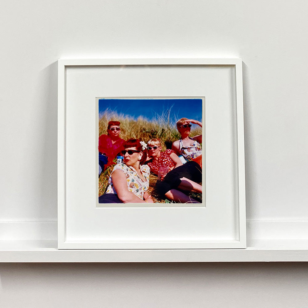 White framed photograph by Richard Heeps. A photo of four women dressed in a retro style all wearing sunglasses on a sunny day lounging on a hill with high grass.