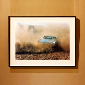Black framed photograph by Richard Heeps. A back view of a light blue Buick car moving and slightly obscured by the dust clouds which it has created.