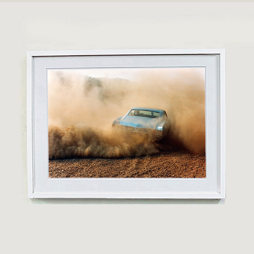 White framed photograph by Richard Heeps. A back view of a light blue Buick car moving and slightly obscured by the dust clouds which it has created.