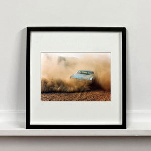 Black framed photograph by Richard Heeps. A back view of a light blue Buick car moving and slightly obscured by the dust clouds which it has created.