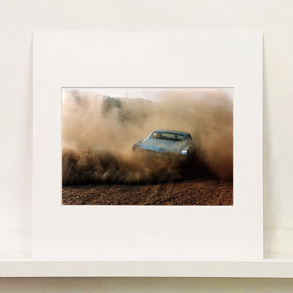 Mounted photograph by Richard Heeps. A back view of a light blue Buick car moving and slightly obscured by the dust clouds which it has created.