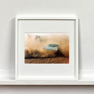 White framed photograph by Richard Heeps. A back view of a light blue Buick car moving and slightly obscured by the dust clouds which it has created.