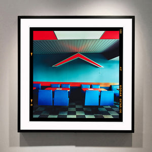 Black framed photograph by Richard Heeps. This photograph captures the inside of a Wimpy Restaurant in Norfolk. There is bright blue seats and red tables. The walls are blue and there is a big red chevron light attached to the wall. 