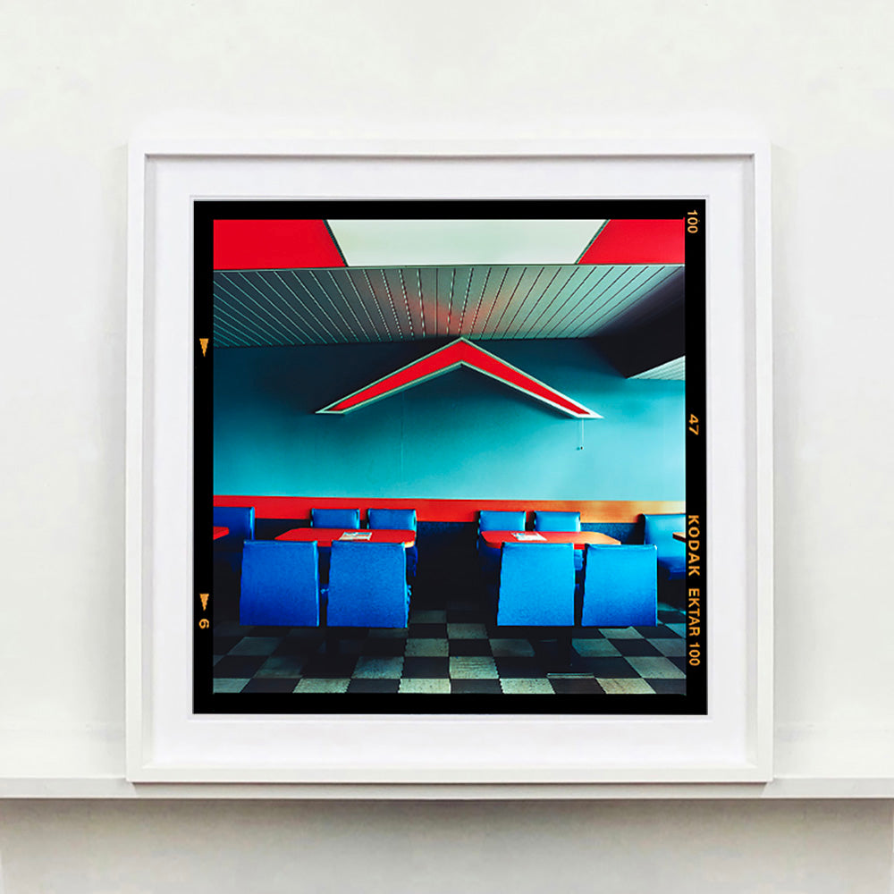 White framed photograph by Richard Heeps. This photograph captures the inside of a Wimpy Restaurant in Norfolk. There is bright blue seats and red tables. The walls are blue and there is a big red chevron light attached to the wall. 