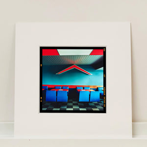 Mounted photograph by Richard Heeps. This photograph captures the inside of a Wimpy Restaurant in Norfolk. There is bright blue seats and red tables. The walls are blue and there is a big red chevron light attached to the wall. 