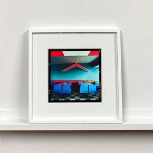 White framed photograph by Richard Heeps. This photograph captures the inside of a Wimpy Restaurant in Norfolk. There is bright blue seats and red tables. The walls are blue and there is a big red chevron light attached to the wall. 
