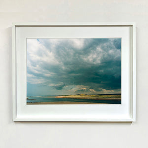 White framed photograph by Richard Heeps. A dark rolling sky sits over this headland pouring different lights over the sea, sand and dunes below.