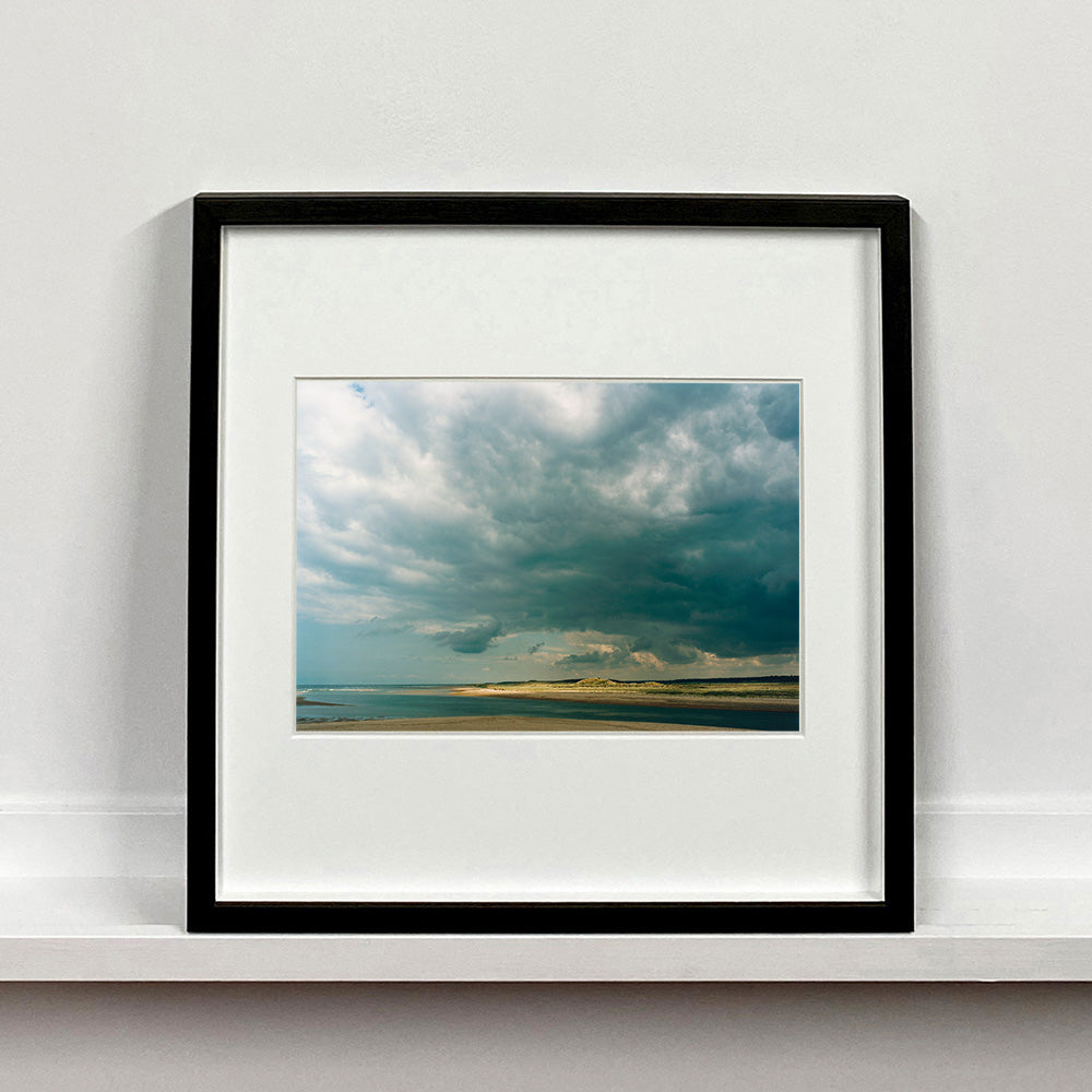Black framed photograph by Richard Heeps. A dark rolling sky sits over this headland pouring different lights over the sea, sand and dunes below.