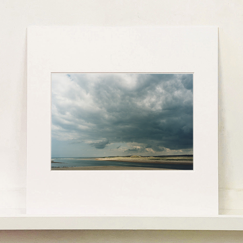 Mounted photograph by Richard Heeps. A dark rolling sky sits over this headland pouring different lights over the sea, sand and dunes below.