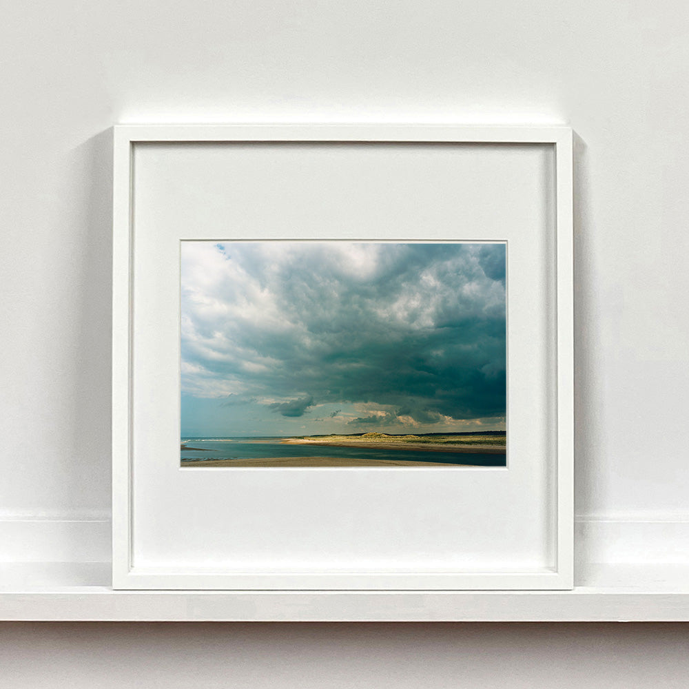White framed photograph by Richard Heeps. A dark rolling sky sits over this headland pouring different lights over the sea, sand and dunes below.