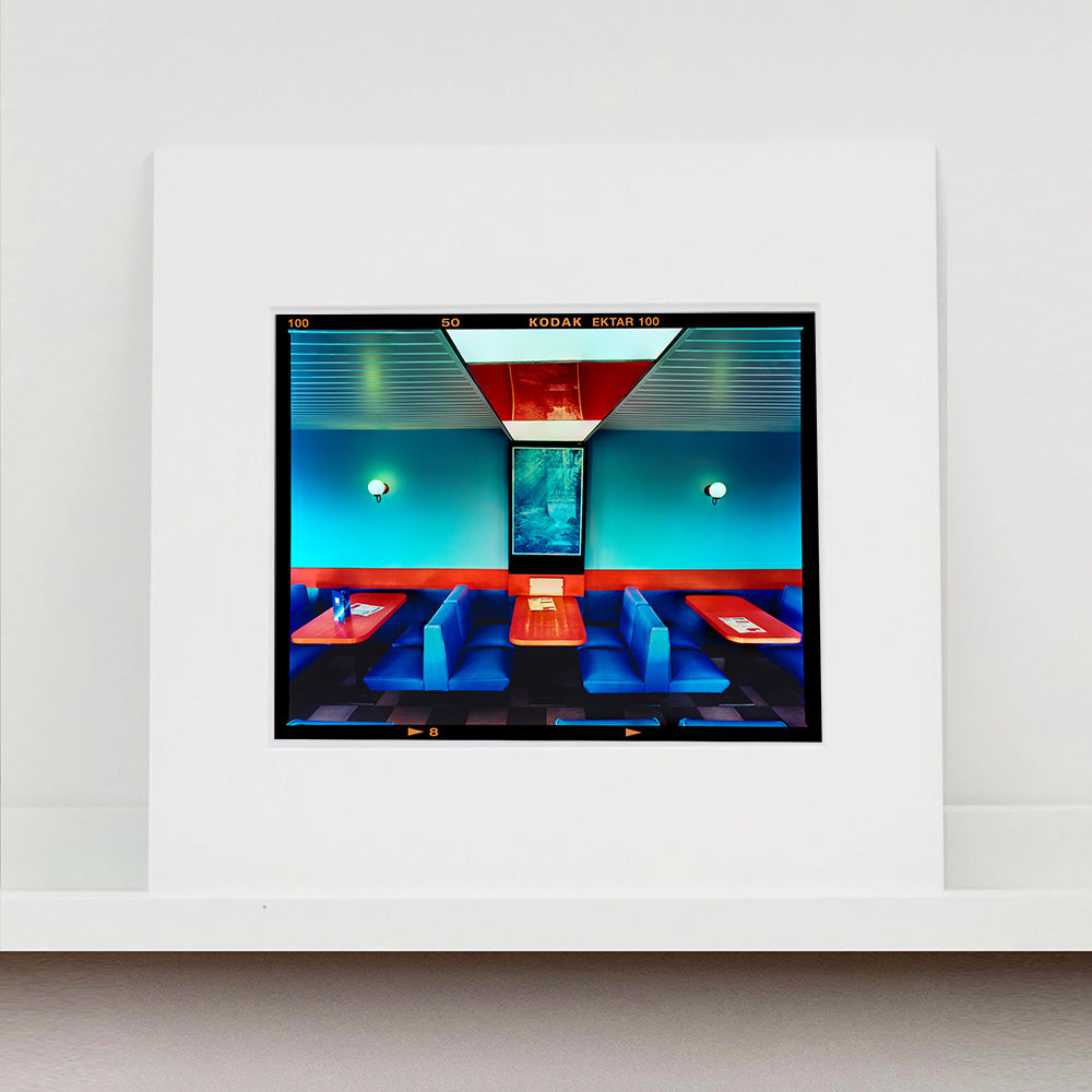 Photograph by Richard Heeps. The inside of a Wimpy restaurant. Blue upholstered double seats pinned to the floor and separated by striking red tables. The walls are blue with a blue picture of trees with a black surround aligned with the middle table.