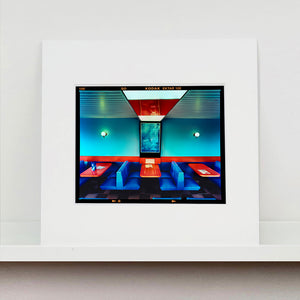 Mounted photograph by Richard Heeps. The inside of a Wimpy restaurant. Blue upholstered double seats pinned to the floor and separated by striking red tables. The walls are blue with a blue picture of trees with a black surround aligned with the middle table.