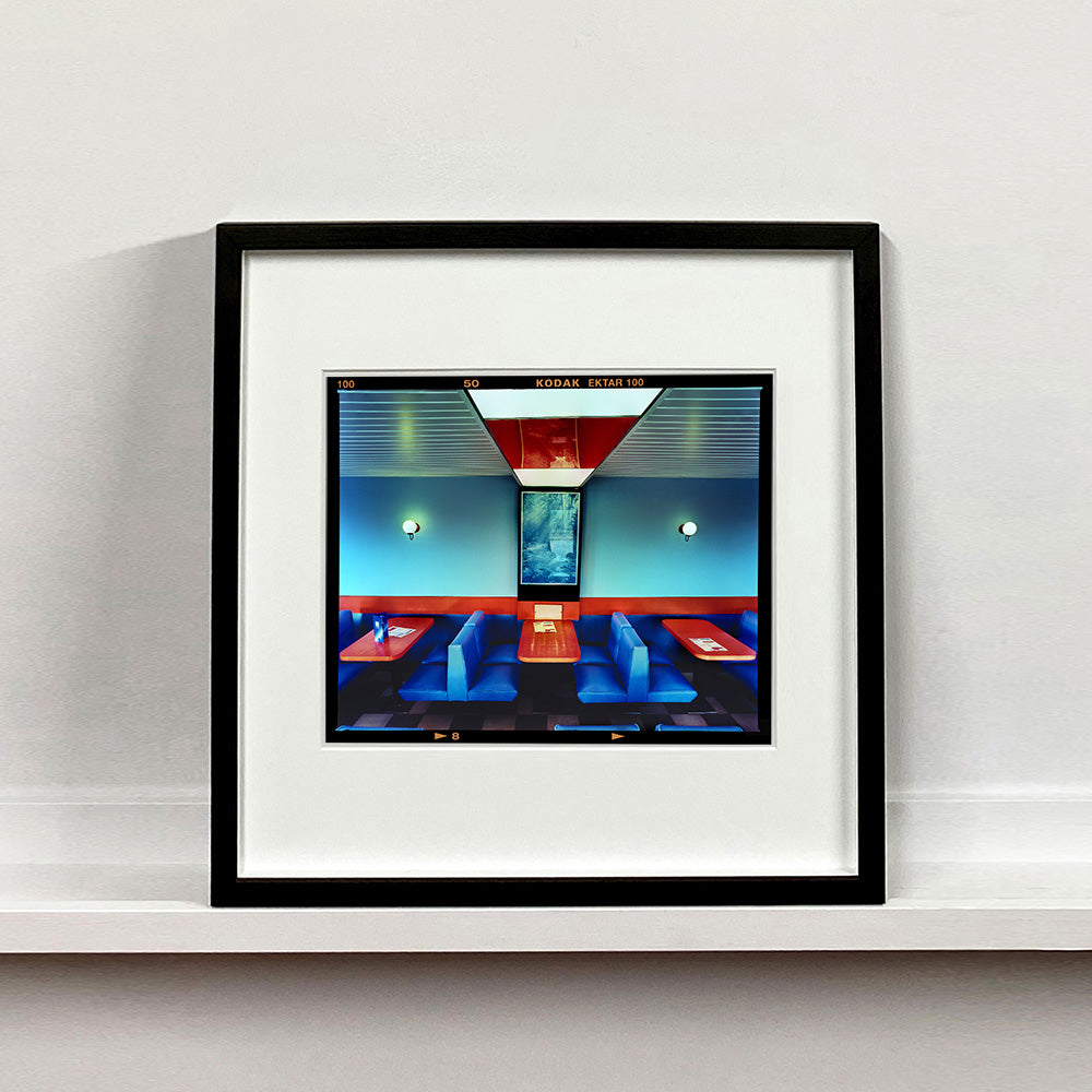 Black framed photograph by Richard Heeps. The inside of a Wimpy restaurant. Blue upholstered double seats pinned to the floor and separated by striking red tables. The walls are blue with a blue picture of trees with a black surround aligned with the middle table.