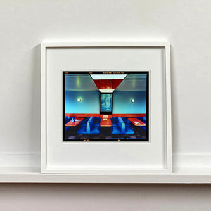 White framed photograph by Richard Heeps. The inside of a Wimpy restaurant. Blue upholstered double seats pinned to the floor and separated by striking red tables. The walls are blue with a blue picture of trees with a black surround aligned with the middle table.