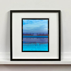 Black framed photograph by Richard Heeps. The blue bay at Llandudno, cut across the middle with path and railings with a gap right in the middle.