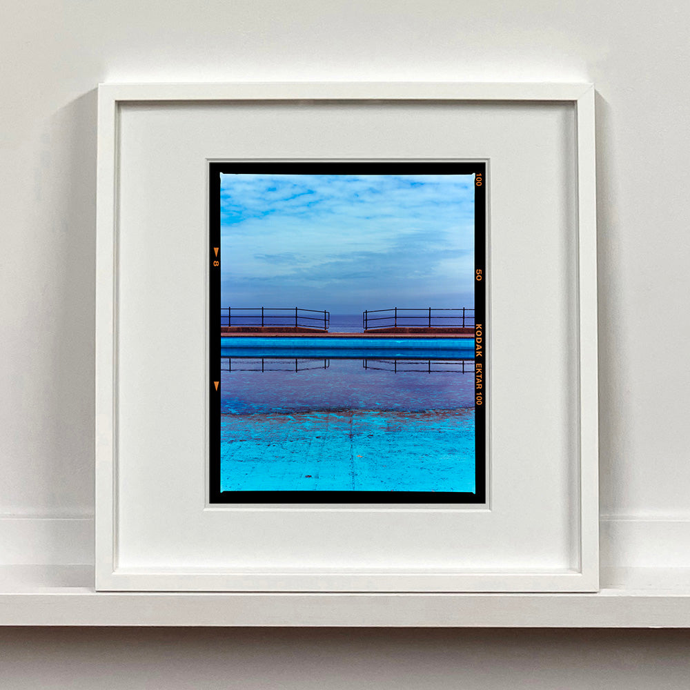 White framed photograph by Richard Heeps. The blue bay at Llandudno, cut across the middle with path and railings with a gap right in the middle.